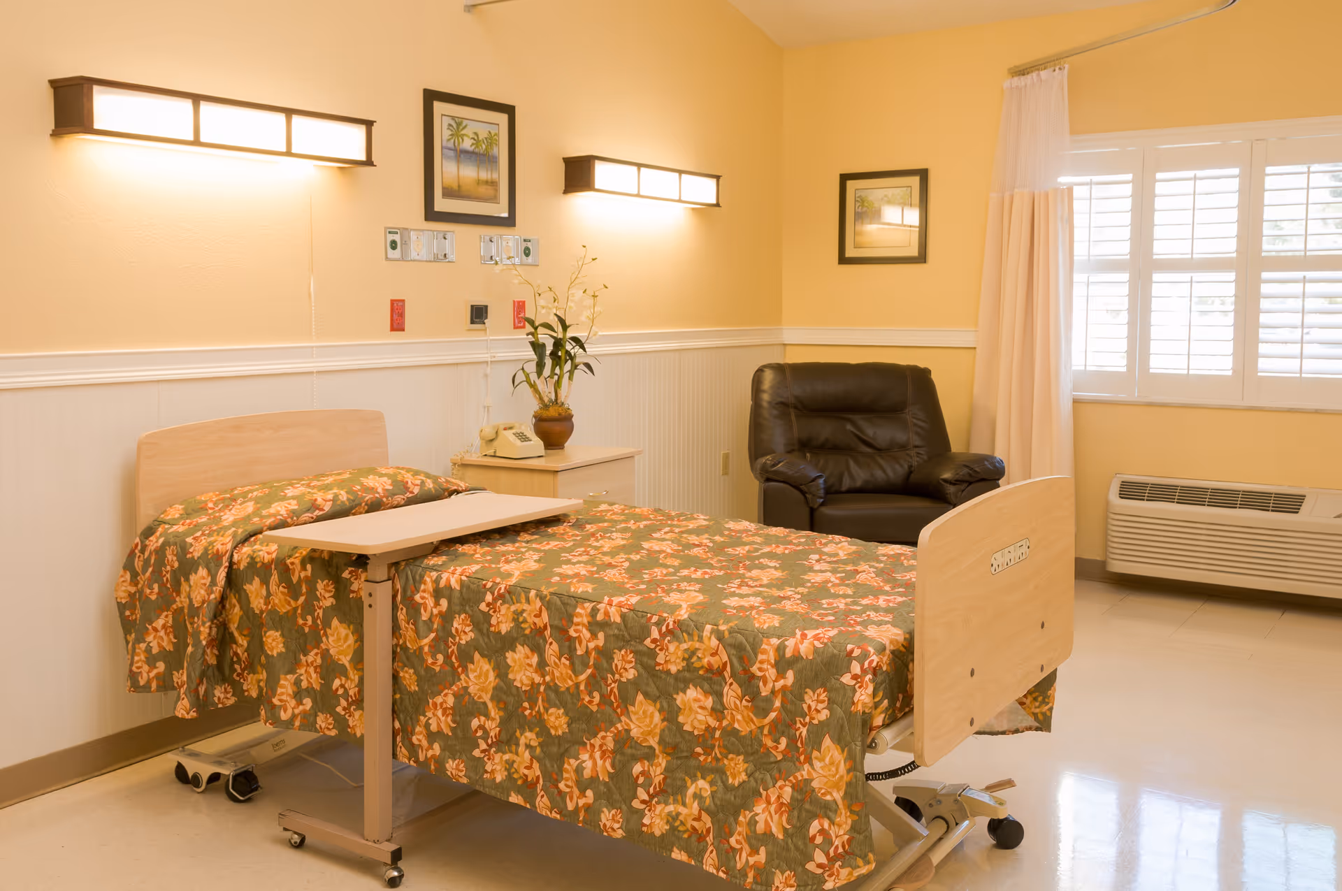 A senior living facility bedroom with a hospital-style bed covered in a floral bedspread, a small overbed table, a nightstand with a telephone and a potted plant, a black leather recliner chair, two framed pictures on the walls, wall-mounted lights, and a window with white shutters and curtains.