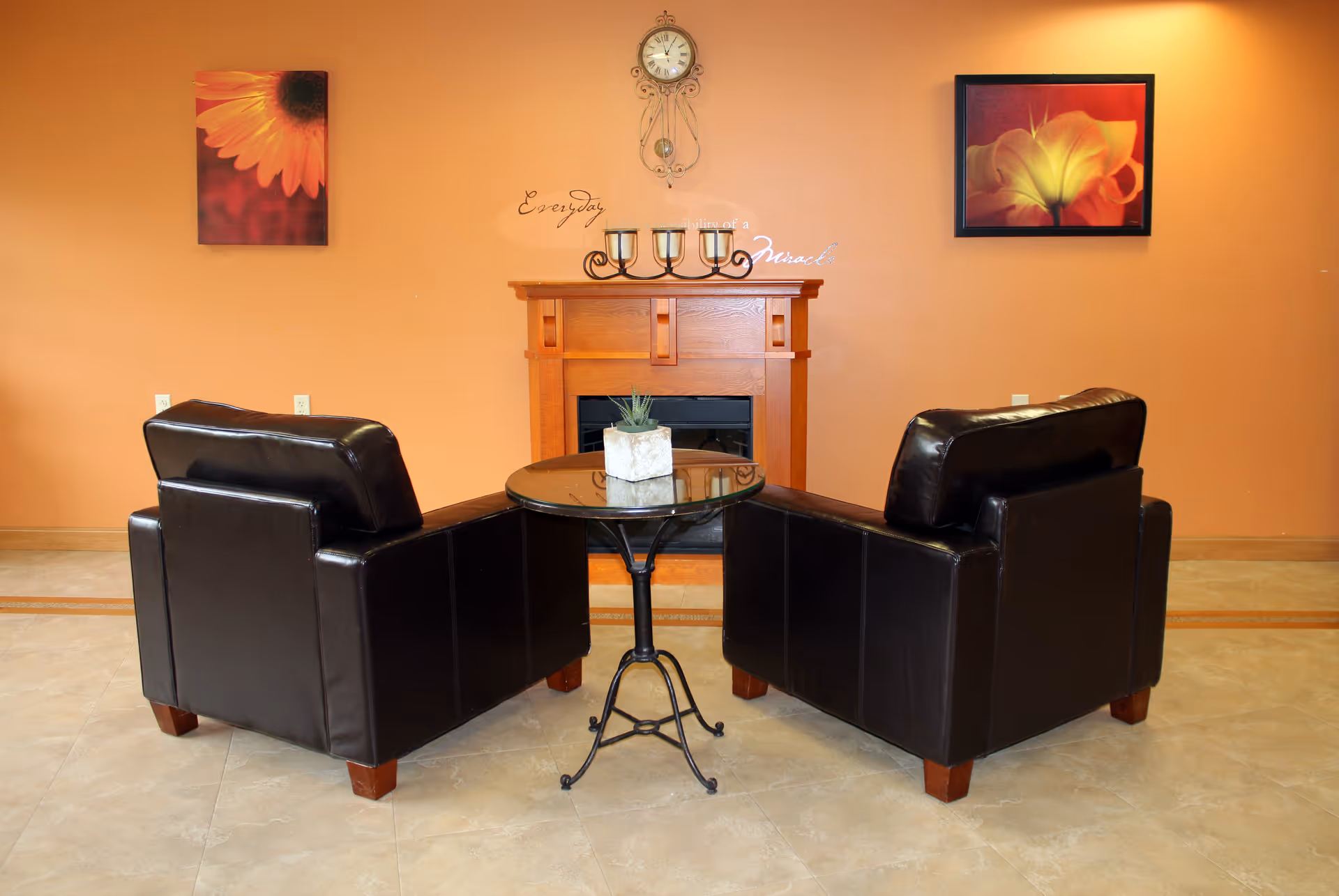 A cozy seating area with two black leather armchairs facing a small round glass table with a potted plant on it. Behind the chairs is a wooden fireplace mantel with three candle holders and a decorative clock mounted on the wall above. The walls are painted a warm orange color and decorated with two framed floral paintings.