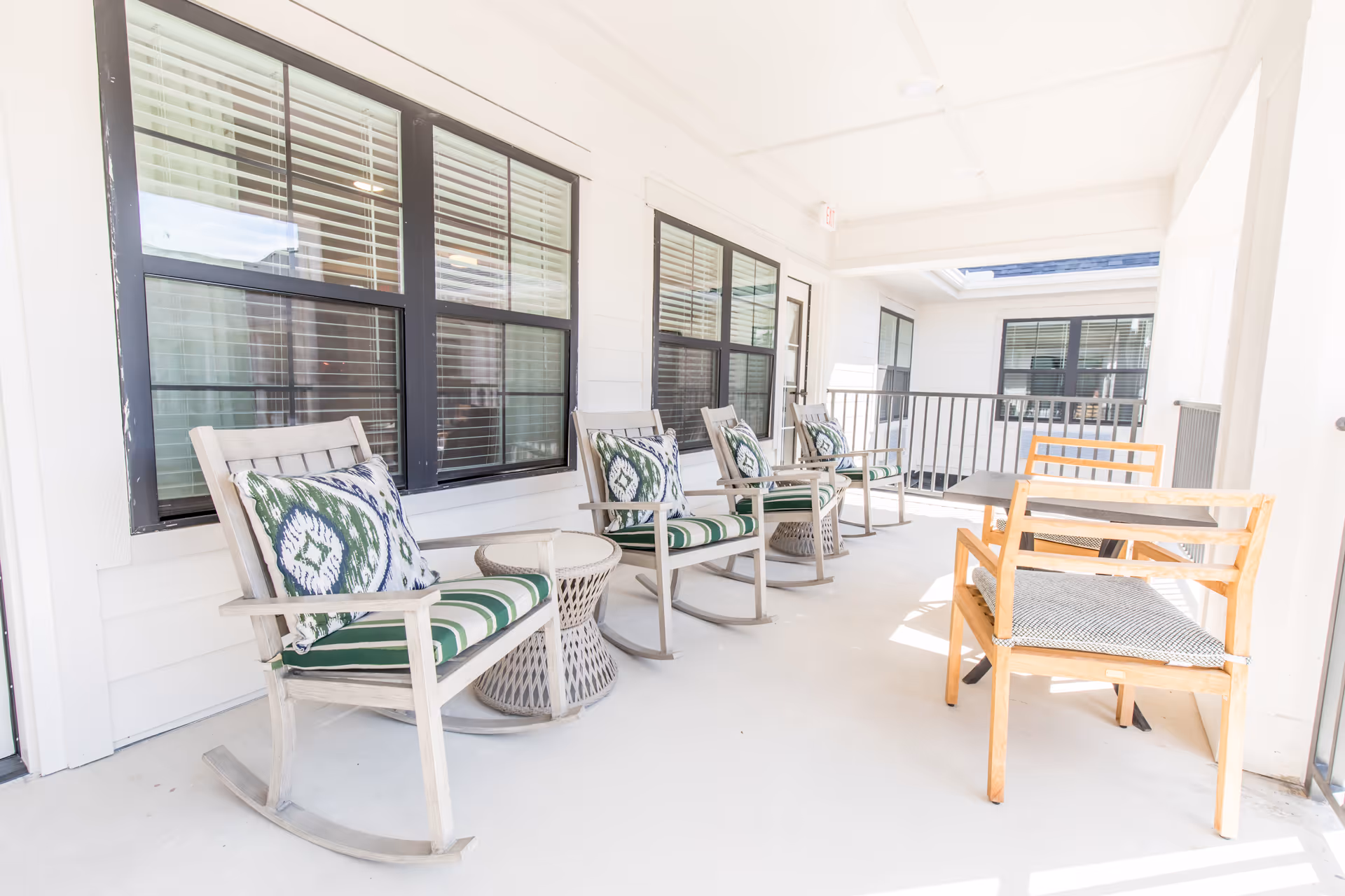 A bright covered outdoor patio area with four wooden rocking chairs featuring green and white patterned cushions and pillows, two small round side tables, and a wooden bench with a cushion next to a rectangular table. The patio is adjacent to a building with white siding and large windows with black frames and blinds.