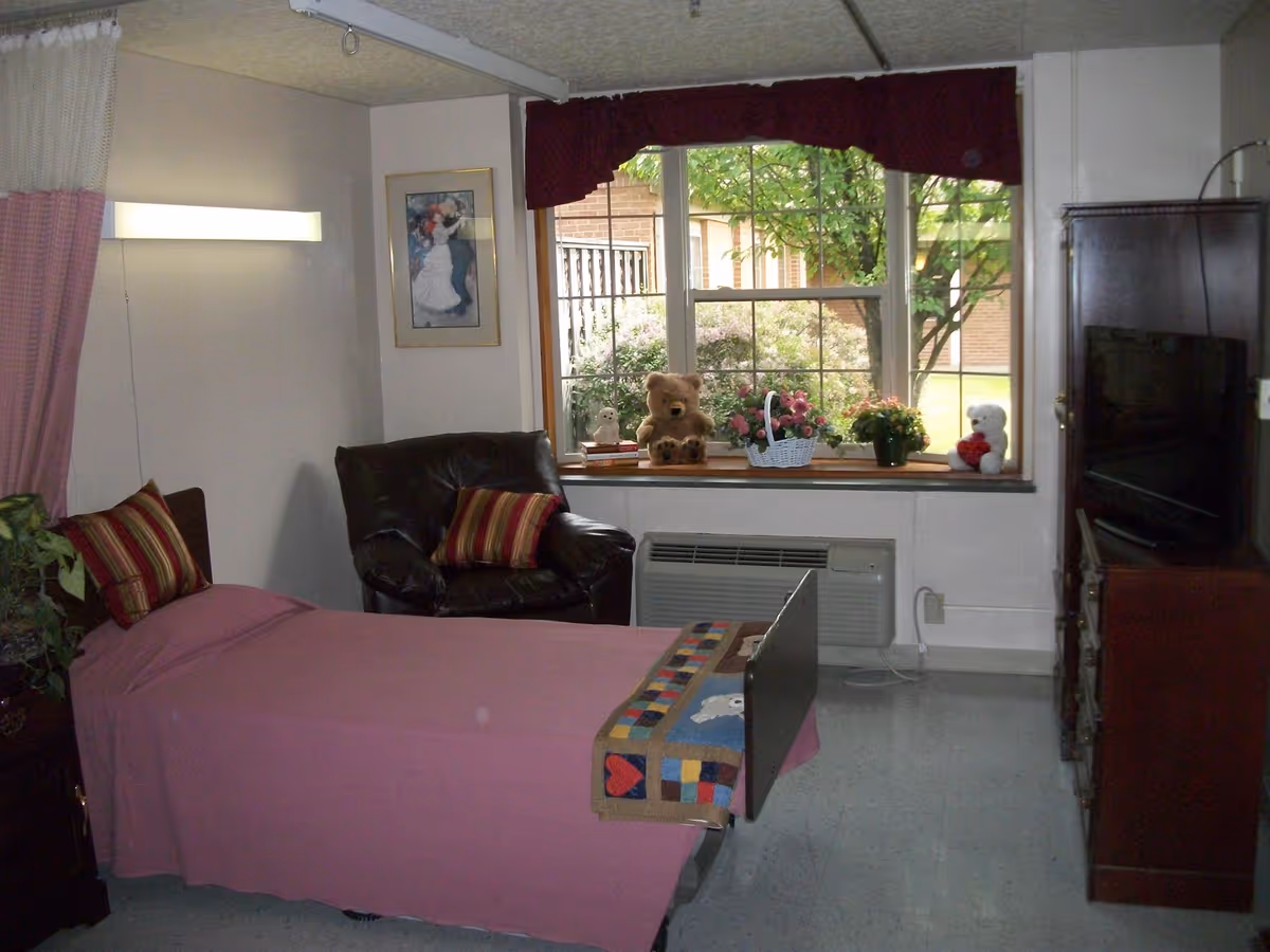 A senior living bedroom with a single bed covered in a pink blanket, a leather armchair, a window sill displaying stuffed animals and flowers, and a TV cabinet.