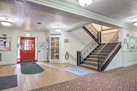 Interior view of a senior living facility lobby with a red entrance door, tiled floor near the entrance, carpeted floor further inside, a staircase with black metal railing leading to the upper floor, and bulletin boards on the walls.