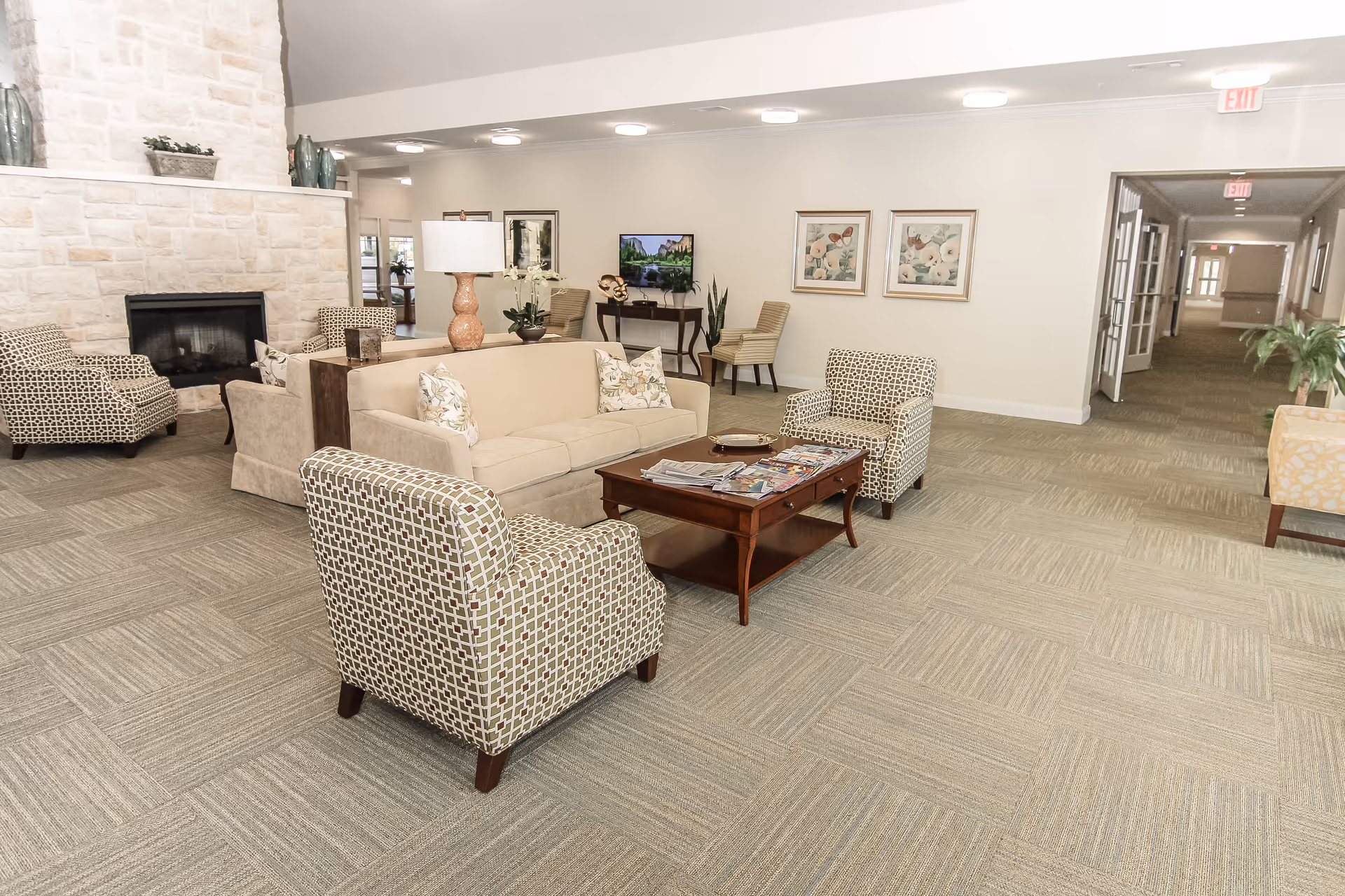 A spacious and well-lit living room area with a beige sofa, two patterned armchairs, a wooden coffee table with magazines, a stone fireplace, and decorative plants and artwork on the walls. The room has a neutral color palette and a carpeted floor, with a hallway visible in the background.