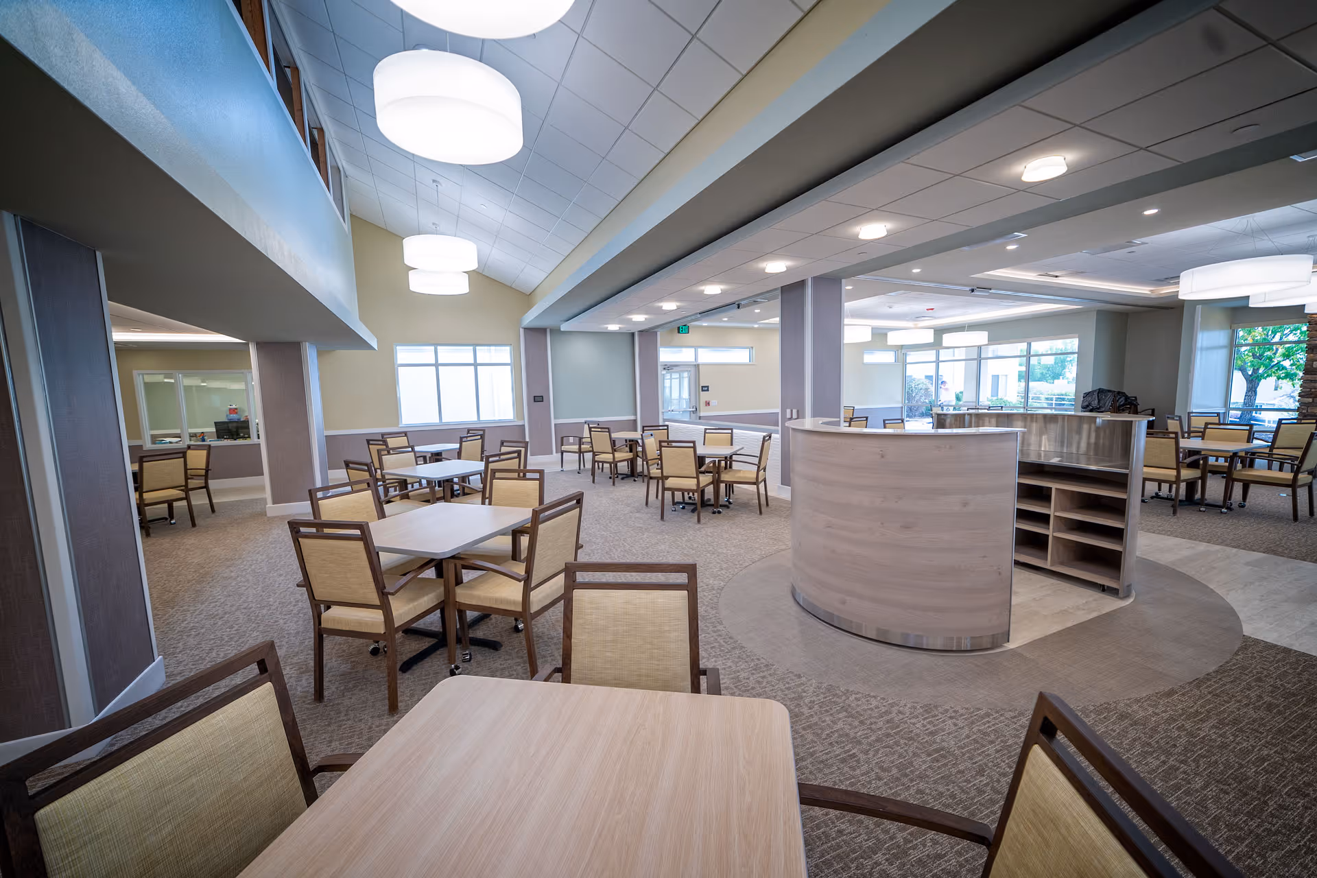 Spacious senior living dining room with multiple tables and chairs, a central service counter, and large pendant lights.
