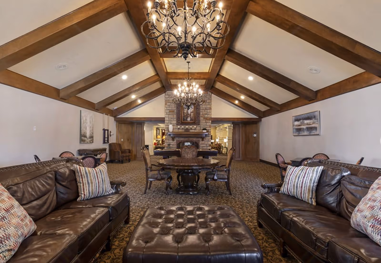 Spacious communal lounge featuring leather sofas, a tufted ottoman, round dining table, stone fireplace, and exposed wooden-beam vaulted ceiling with chandeliers.
