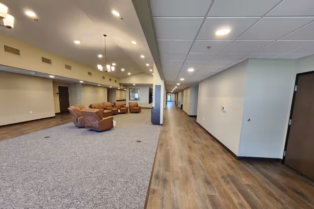 Spacious memory care common area with brown recliners clustered on carpet to the left and a long wood-floored corridor to the right.
