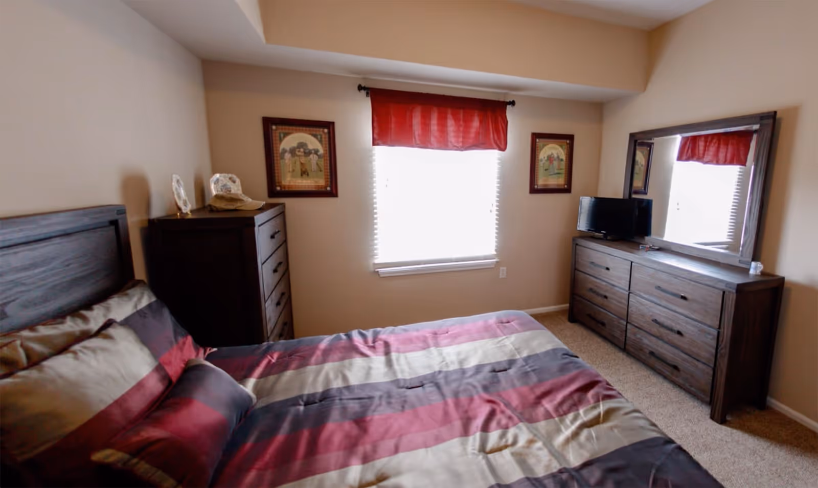 A bedroom with a bed covered in a striped comforter in shades of red, gray, and beige. The room has a window with white blinds and a red valance. On either side of the window are two framed pictures. There is a dark wooden dresser with a mirror on the right side of the room, topped with a small TV. On the left side, there is a tall dark wooden chest of drawers with decorative items on top. The walls are painted beige and the floor is carpeted.