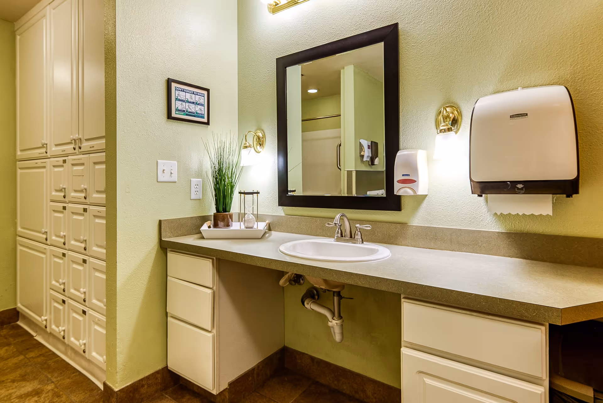 A clean and well-lit bathroom area with a countertop sink, a large mirror with a dark frame, wall-mounted soap dispenser and paper towel holder, two wall sconces with lights, and white cabinets underneath the counter and along the adjacent wall. A small decorative plant and an hourglass are placed on the countertop.
