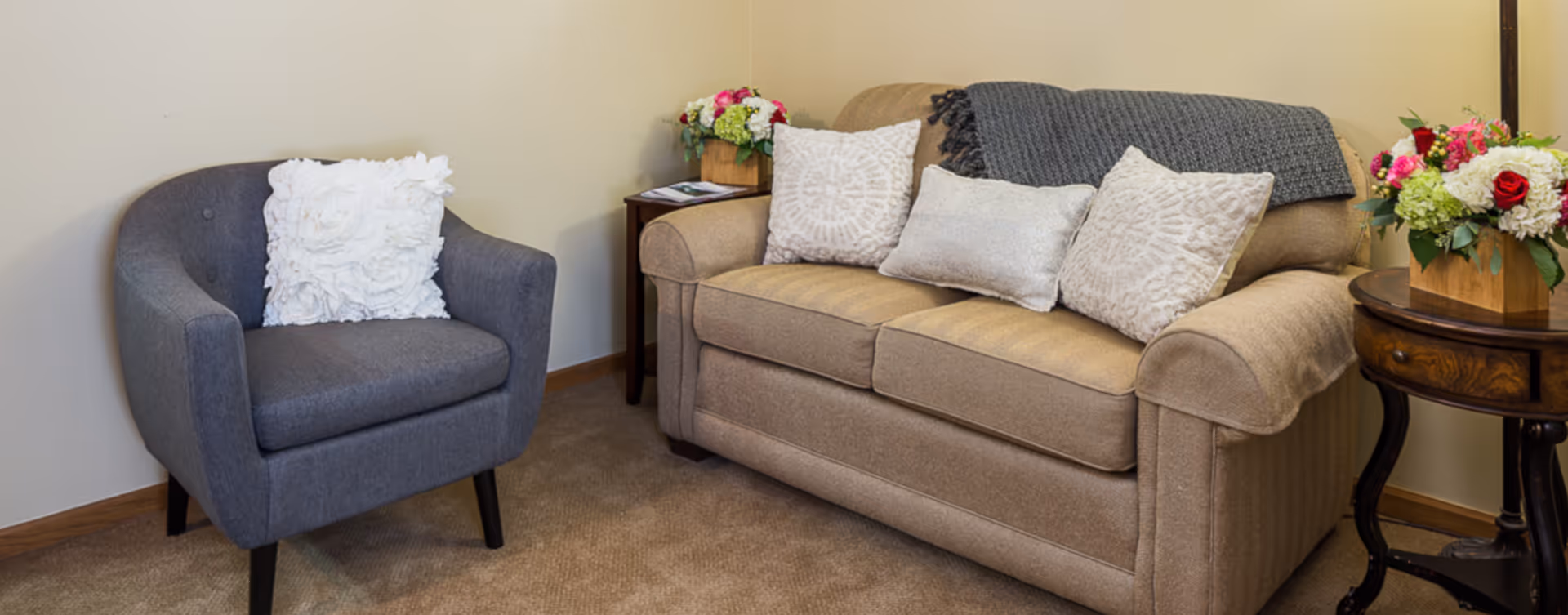 A cozy living room area featuring a beige sofa with three decorative pillows and a gray throw blanket, a gray armchair with a white textured pillow, two wooden side tables each holding a floral arrangement, and beige carpeted flooring.