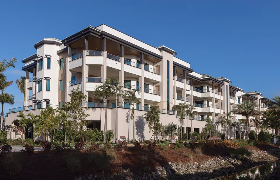 Exterior view of a modern multi-story senior living facility building with balconies, surrounded by palm trees and landscaping under a clear blue sky.