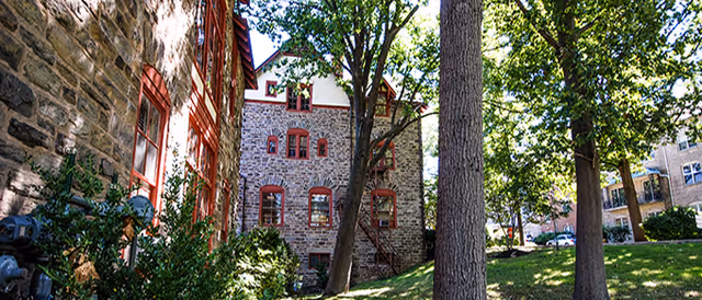 Exterior view of a senior living community building made of stone with red window frames, surrounded by large trees and green grass on a sunny day.