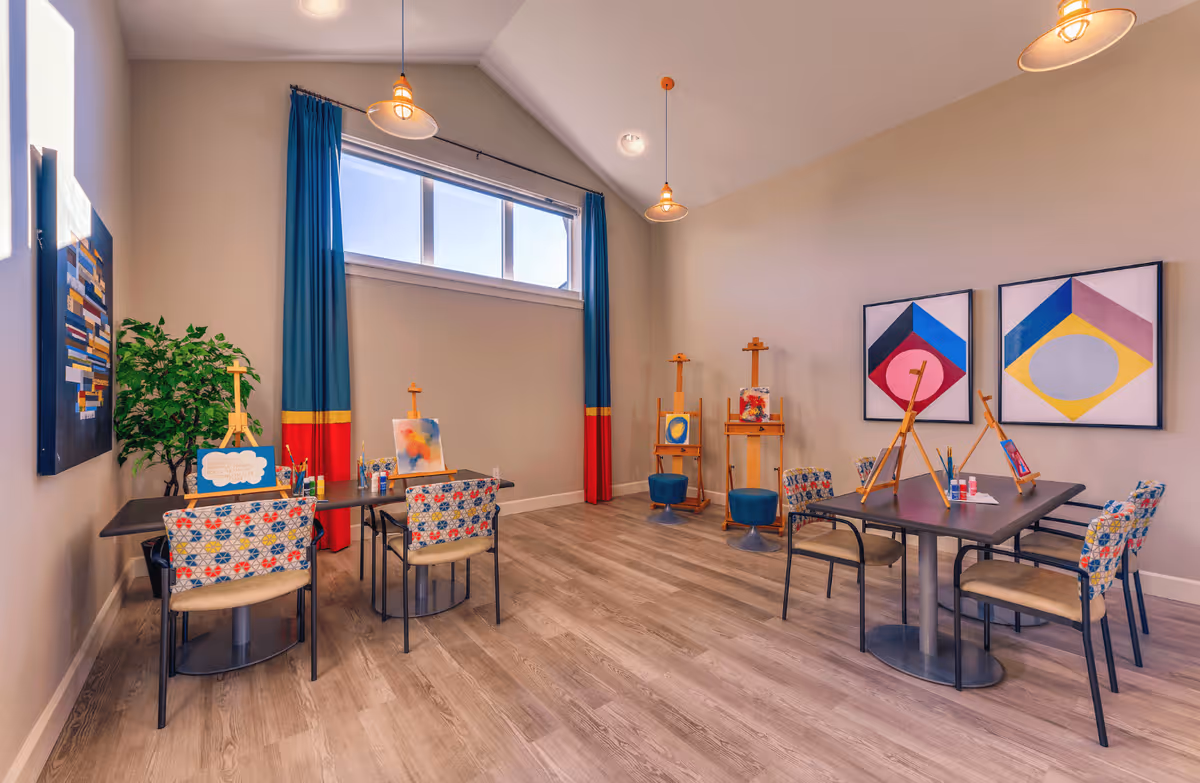 A bright art room with two tables and colorful patterned chairs arranged around them. Each table has small easels with paintings and art supplies. The room features large windows with blue and red curtains, wooden flooring, and modern pendant lights hanging from a high ceiling. Abstract artwork is displayed on the walls.