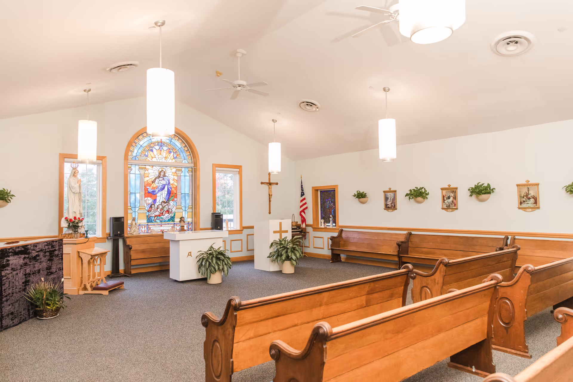 Interior of a small chapel with wooden pews, a stained glass window depicting a religious figure, a statue, an altar, and religious decorations on the walls. The room is well-lit with hanging lights and ceiling fans.