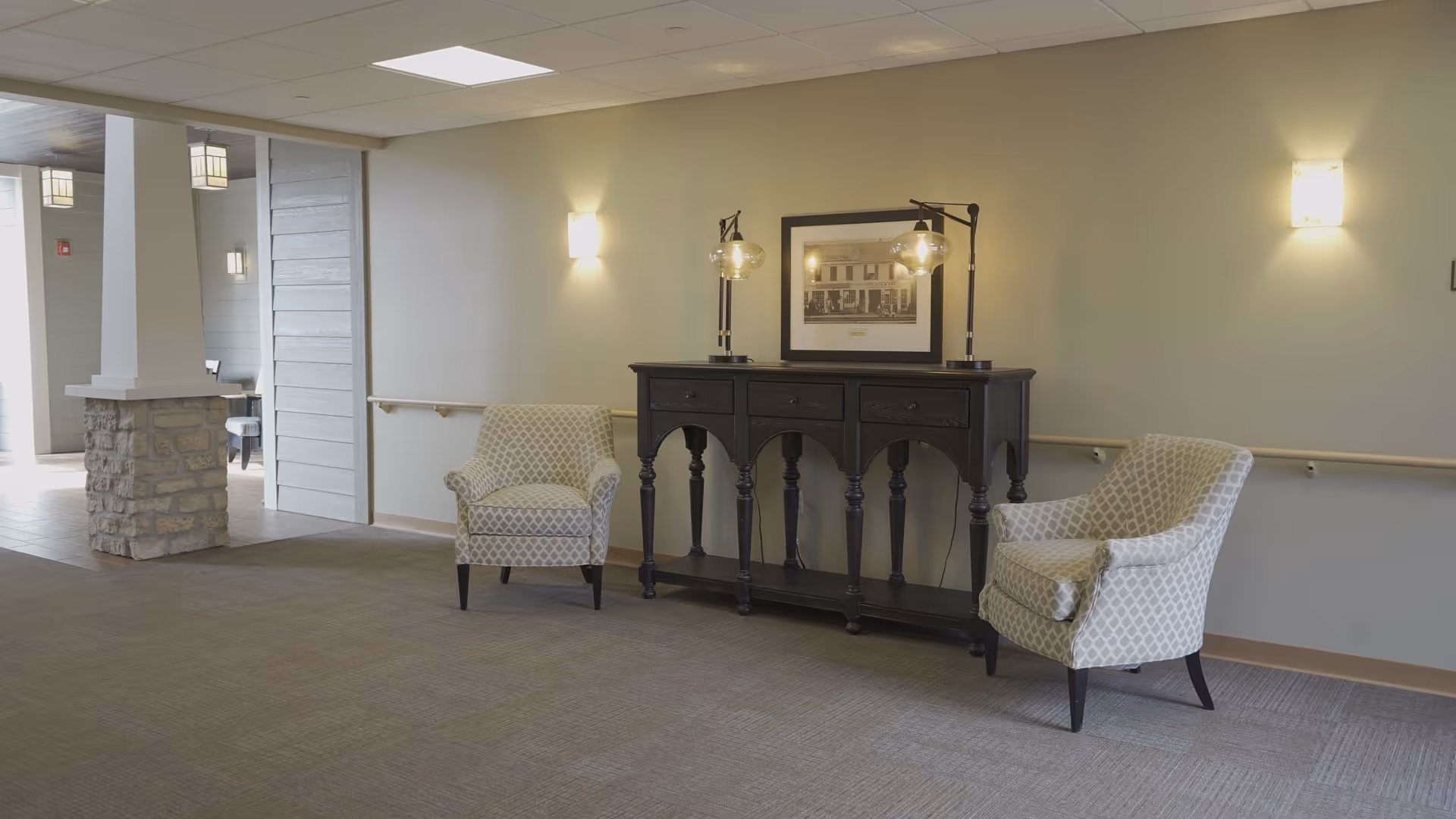 A hallway area in a senior living facility with two patterned armchairs placed on either side of a dark wooden console table with three drawers. The table has two lamps with glass shades and a framed black and white photograph hanging above it. The walls are light-colored with wall-mounted lights, and there is a handrail along the wall. The floor is carpeted, and there is a stone pillar and an open doorway visible on the left side.