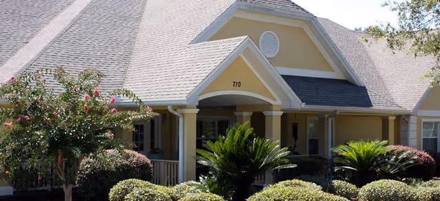 Front exterior view of a single-story assisted living facility building with a beige and cream color scheme, a gabled roof, the number 710 above the entrance, surrounded by well-maintained shrubs and small trees.