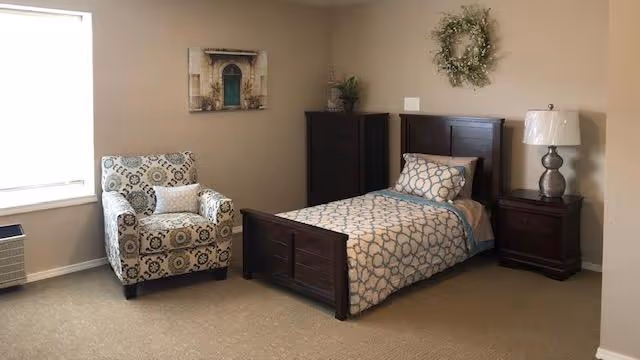 A furnished bedroom with a single wooden bed, nightstand and lamp, patterned armchair, dresser, and wall decor.