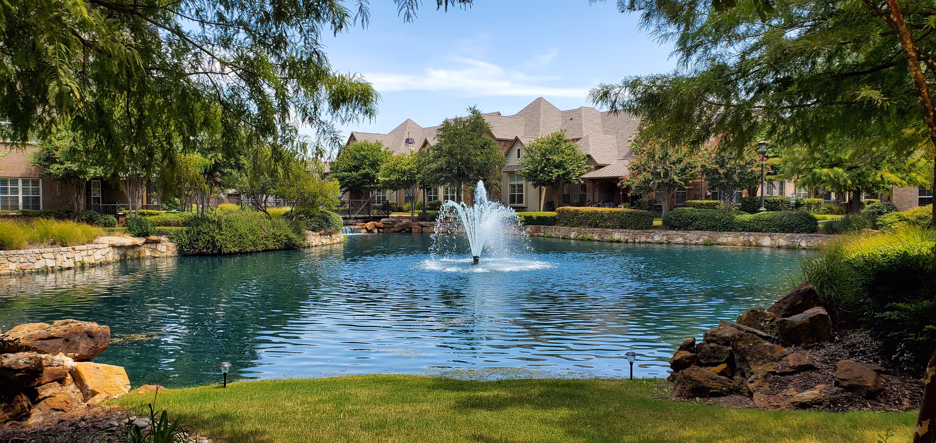 A serene outdoor pond with a central water fountain surrounded by lush greenery and trees, with a large residential building in the background under a clear blue sky.