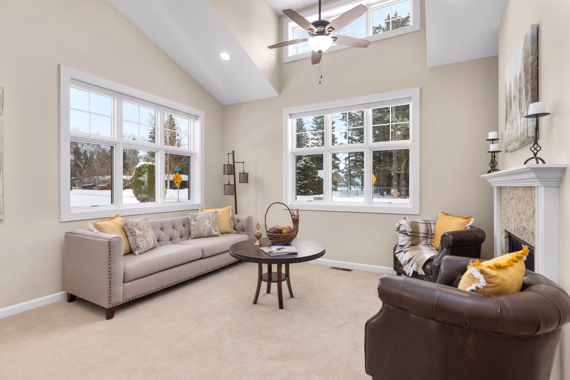 A bright living room with beige walls and carpet, featuring a light gray tufted sofa with patterned and yellow pillows, two dark brown leather armchairs with yellow pillows and a throw blanket, a round wooden coffee table with decorative items, a white fireplace with candles and a painting above it, large windows showing a snowy outdoor scene, and a ceiling fan with lights.
