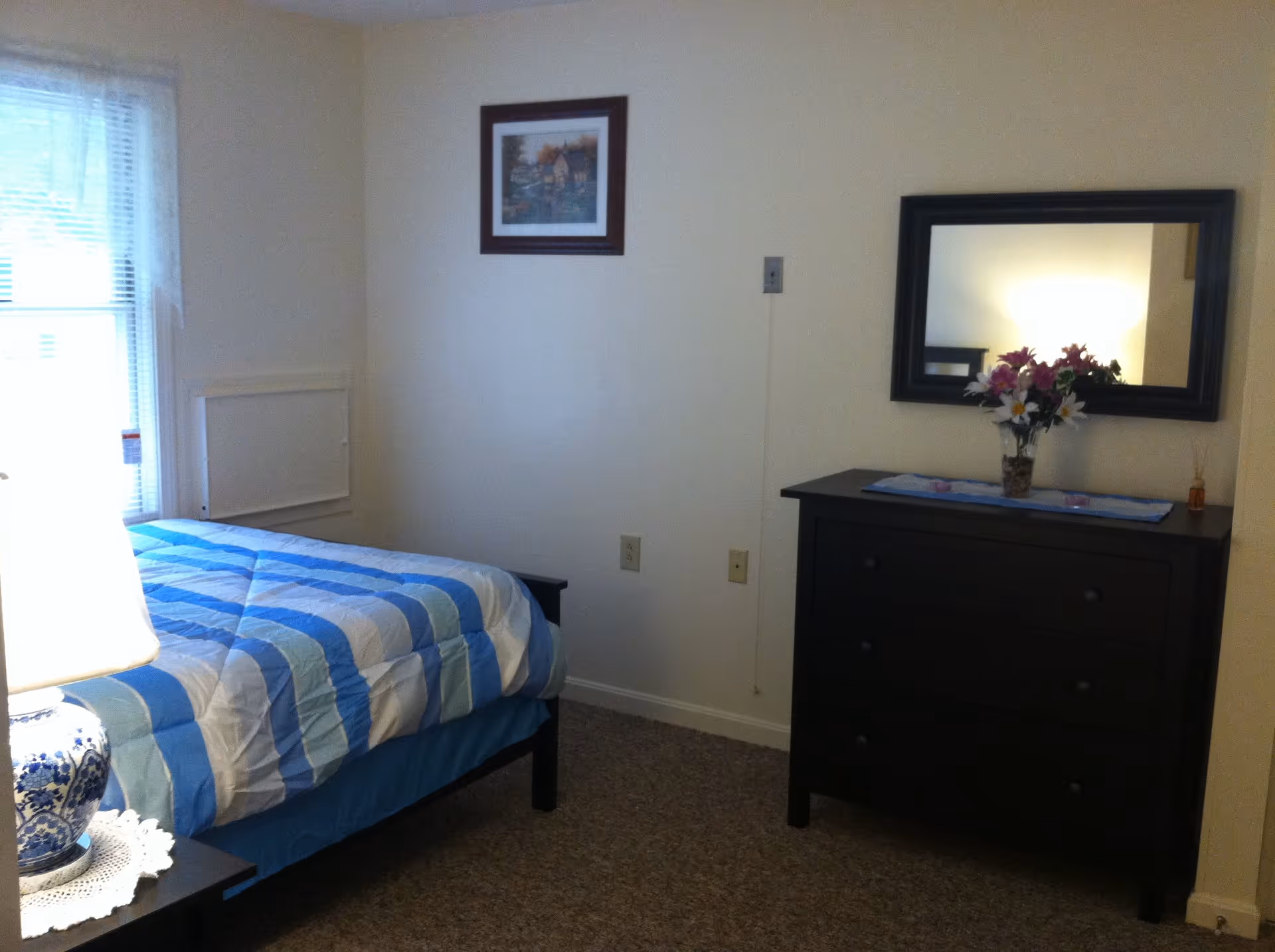 A small bedroom with a single bed covered in a blue and white striped comforter. There is a nightstand with a blue and white lamp and a doily next to the bed. On the opposite wall, there is a dark wooden dresser with three drawers, a vase of flowers on top, and a large rectangular mirror hanging above it. A framed picture is hung on the wall above the bed. The room has beige walls and carpeted floor.