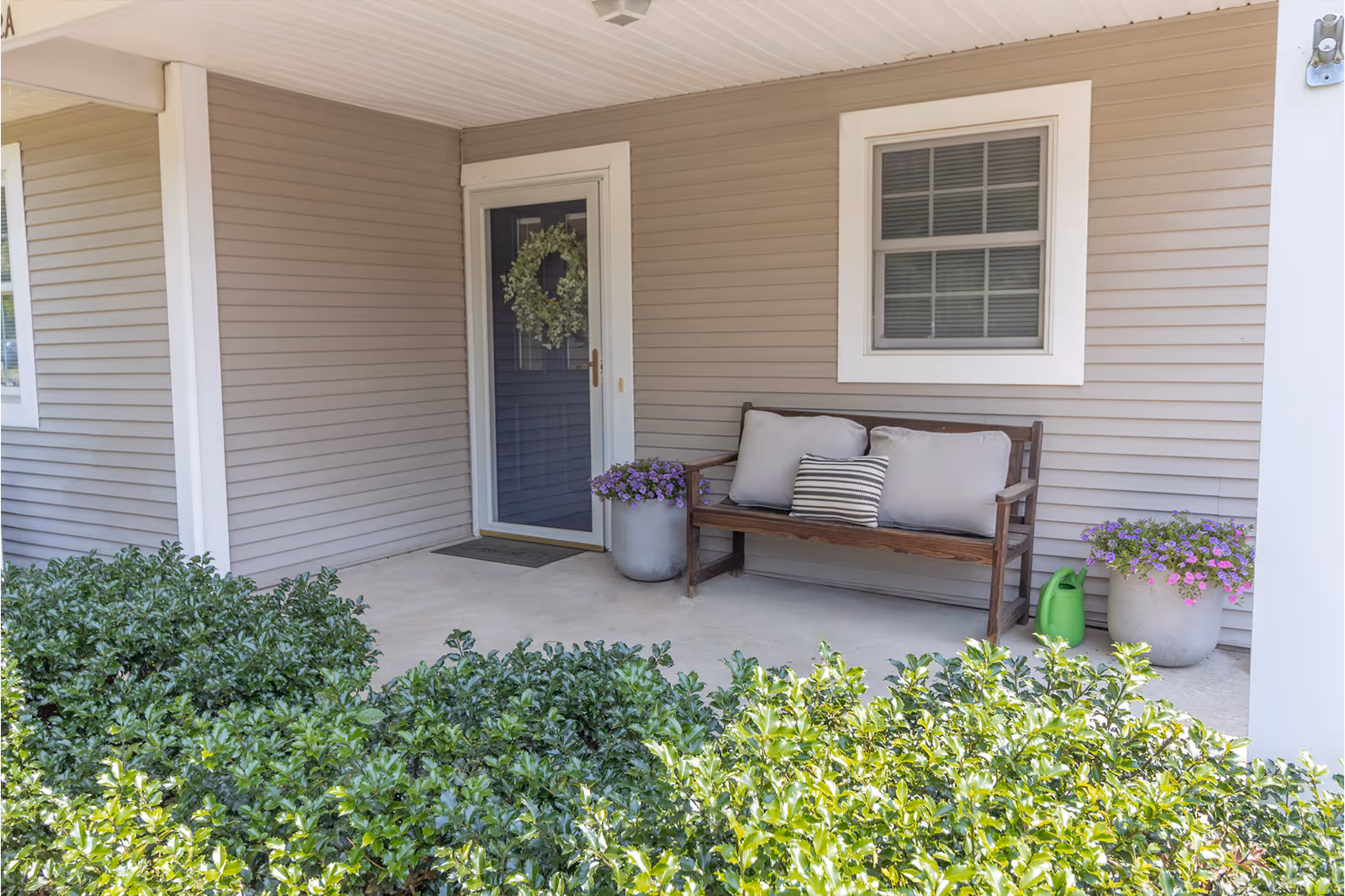 Covered front porch with a wooden bench with cushions, potted flowers, a wreath-adorned door, and shrubs in front.