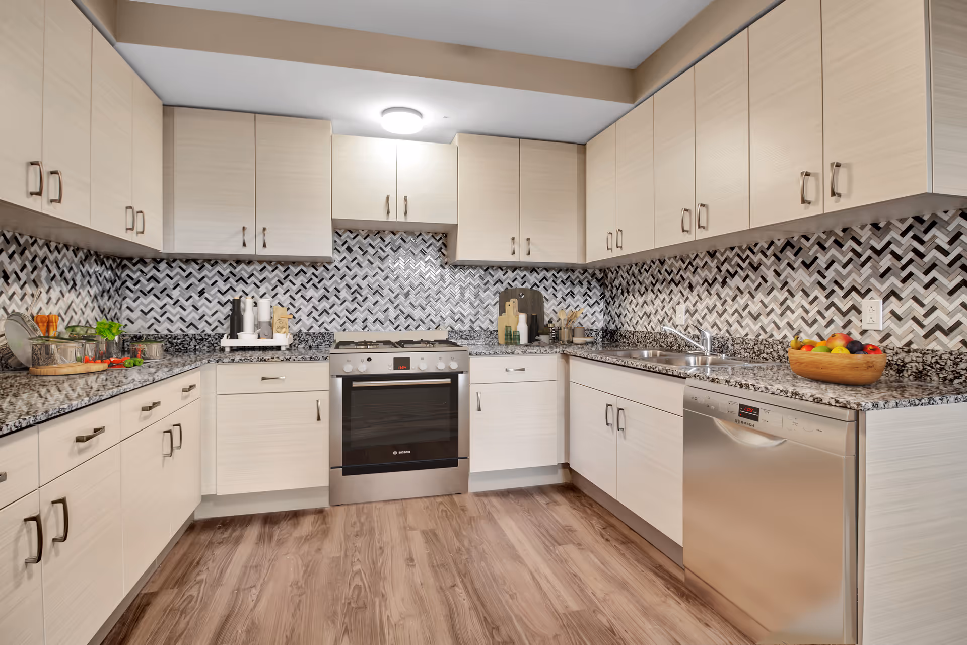 Modern kitchen with light wood cabinets, granite countertops, a stainless steel oven and dishwasher, a patterned black and white tile backsplash, and wood flooring. Various kitchen items and a bowl of fruit are placed on the countertops.
