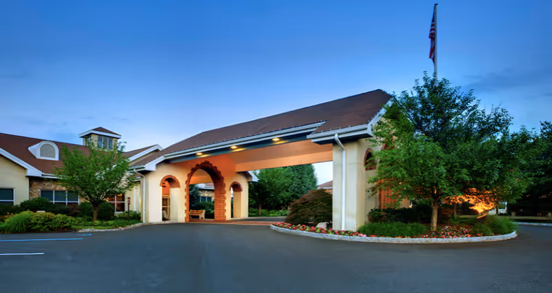 Exterior view of Brittany Pointe Estates senior living facility at dusk, showing a covered entrance with arches, landscaped greenery, and an American flag on a flagpole.