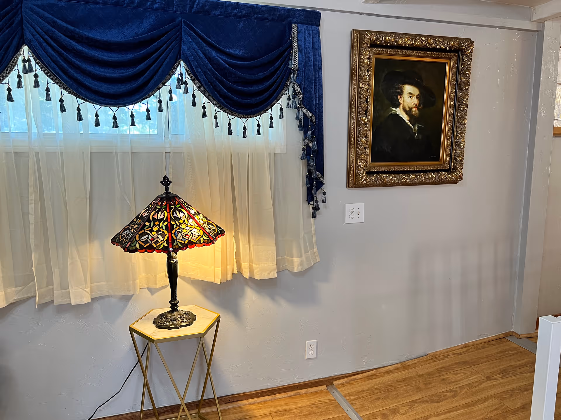 Corner of a living room with a lit stained-glass table lamp on a small side table under a window with a blue valance and a framed portrait on the wall.