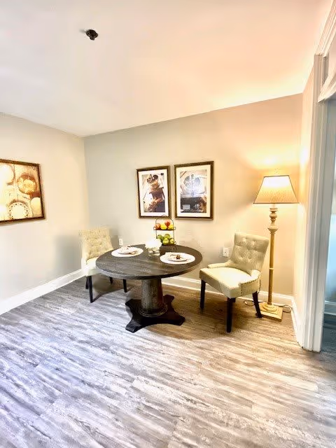 A small dining area with a round dark wooden table set for two with plates and cutlery. Two upholstered chairs with tufted backs are placed on either side of the table. A floor lamp with a beige shade stands to the right, and three framed pictures hang on the beige walls. The floor has light gray wood-like flooring.