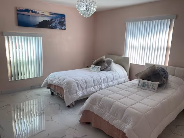 A bright bedroom with two single beds, each covered with white comforters and decorative pillows. One pillow says 'HAPPY' and the other says 'LOVE'. The room has light pink walls, two windows with vertical blinds, a chandelier-style light fixture, and a large landscape photograph hanging on the wall. The floor is white with a marble pattern.