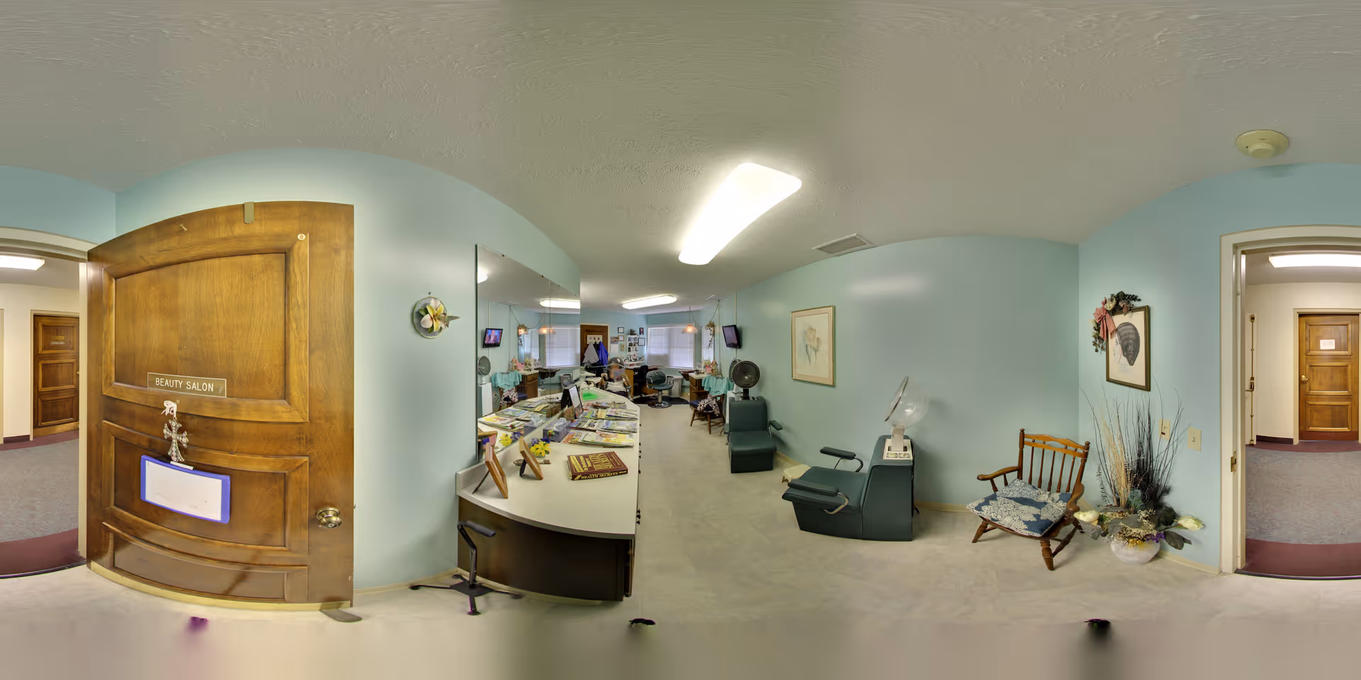 Interior view of a beauty salon in a senior living facility with light blue walls, a wooden door labeled 'Beauty Salon', a counter with magazines and decorations, salon chairs, a rocking chair, and framed artwork on the walls.