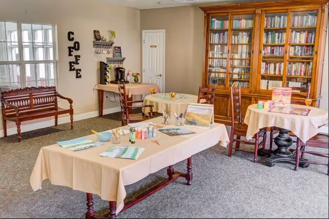 A cozy room with tables covered in beige tablecloths set up for painting activities, including paint bottles, brushes, and canvases. There is a large wooden bookshelf filled with books against the wall, a small coffee station with a coffee maker, and a wooden bench near a window. The word 'COFFEE' is displayed vertically on the wall.