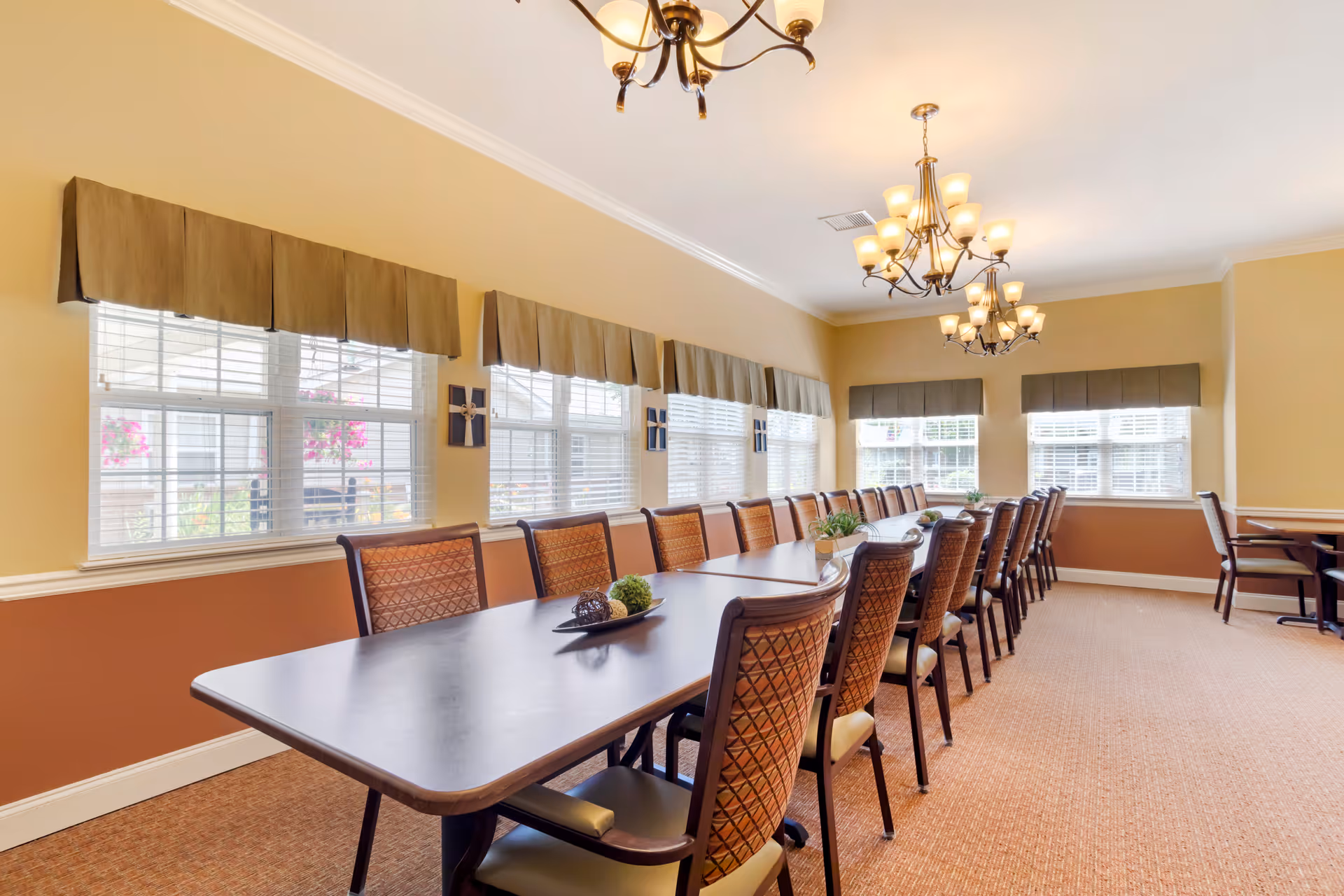 A long dining room with a large rectangular table surrounded by multiple cushioned chairs. The room has beige walls with a darker brown wainscoting, several windows with valances, and two chandeliers hanging from the ceiling. Decorative plants and ornaments are placed on the table.