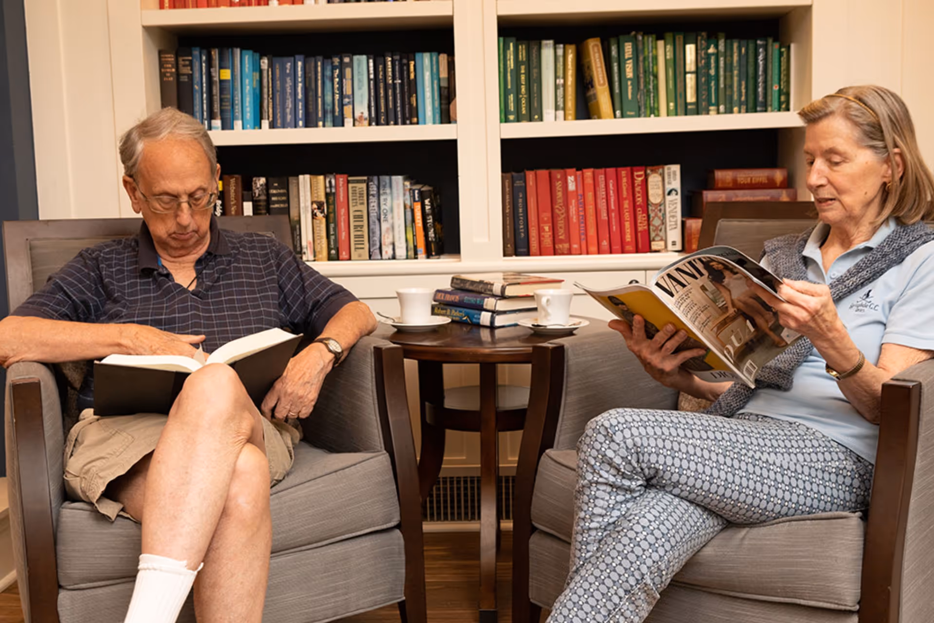 An elderly man and woman sitting in armchairs in a cozy room with bookshelves filled with colorful books behind them. The man is reading a book while the woman is reading a magazine. A small round table between them holds stacked books and two white coffee cups.