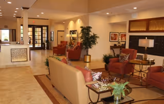 A spacious and well-lit assisted living facility lobby with beige tiled floors, a reception desk on the left, comfortable seating including a beige sofa and red armchairs, decorative plants, framed artwork on the walls, and a glass door entrance in the background.