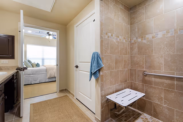 Interior view of a senior living facility bathroom with beige tiled walls and floor. There is a fold-down shower seat and a grab bar on the wall. A blue towel hangs on a hook on a white door. The bathroom opens into a bedroom with a bed, pillows, and a ceiling fan visible.