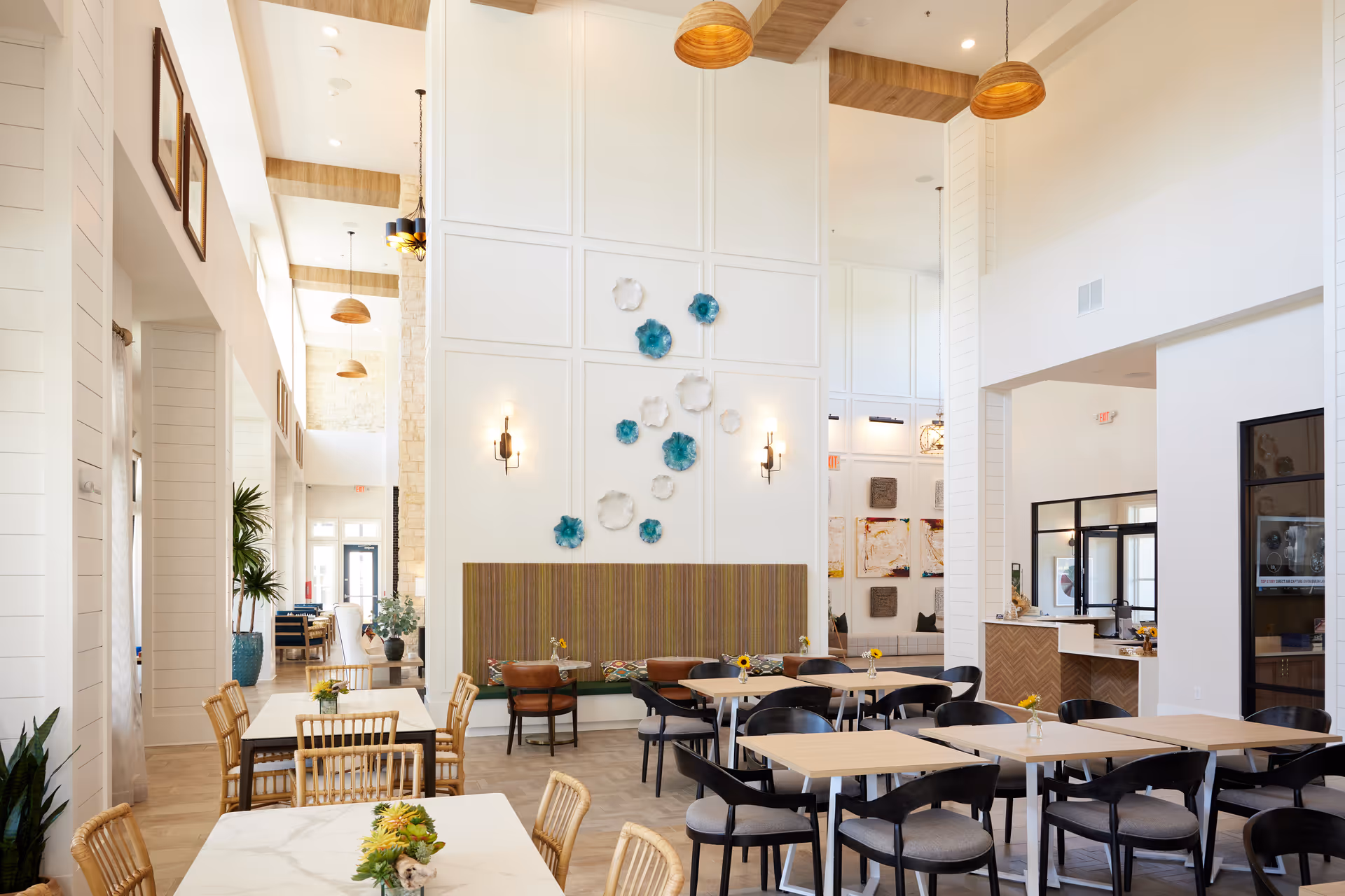 Bright and spacious dining area with high ceilings, featuring multiple tables and chairs arranged for seating. The walls are decorated with blue and white floral wall art, and there are pendant lights hanging from the ceiling. The space has a modern and welcoming design with natural light coming through large windows.