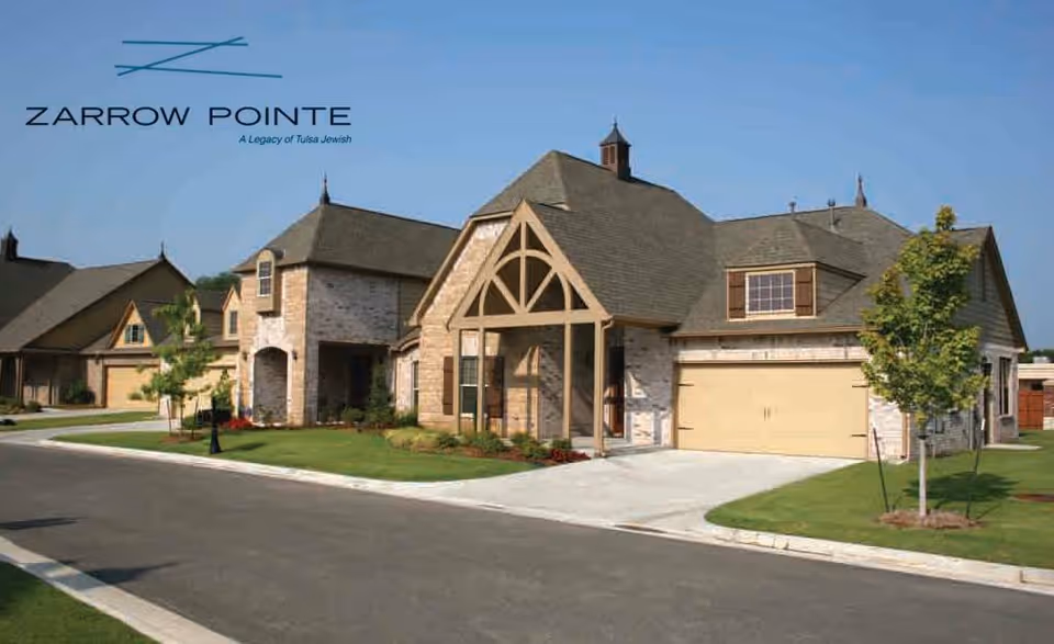 Exterior view of a residential-style building with stone and wood accents, a peaked roof, and a garage. The building is part of a community with well-maintained lawns and a paved road in front. The sky is clear and blue.