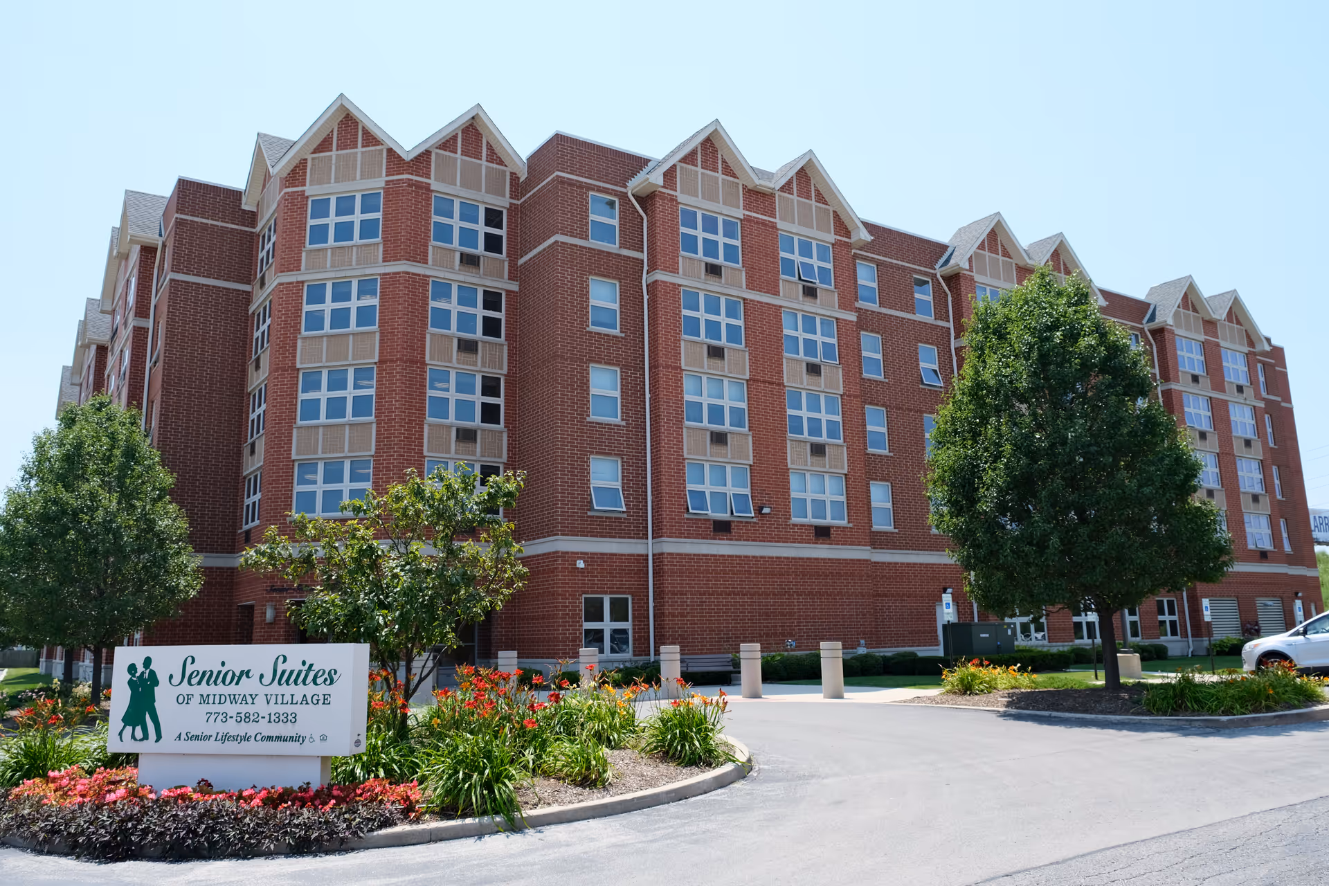 Red-brick, multi-story senior living building with landscaping and a 'Senior Suites of Midway Village' sign in front.