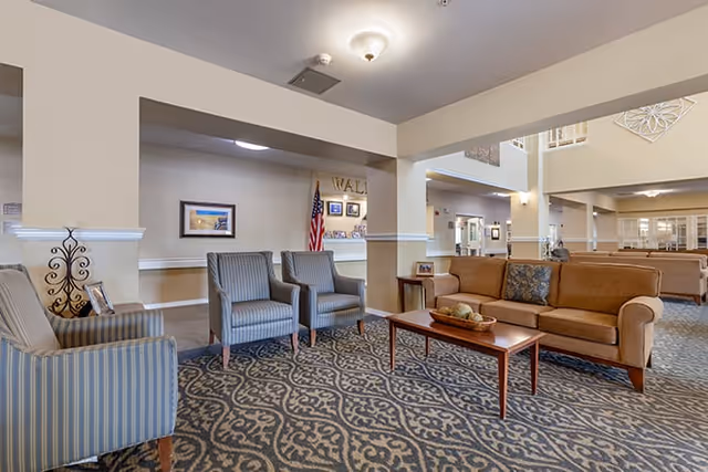 A spacious senior living facility common area with a patterned carpet, beige walls, and ceiling lights. The seating area includes a brown sofa with decorative pillows, two striped armchairs, and a wooden coffee table with a decorative bowl. In the background, there is an American flag, framed pictures on the wall, and a reception desk.