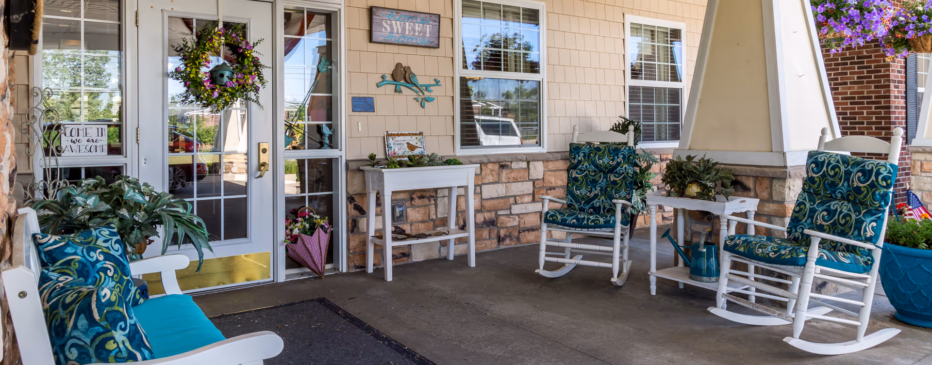 Covered outdoor porch area with white rocking chairs and a bench, all with blue patterned cushions. There are potted plants, a small white table with a watering can, and decorative signs on the wall including one that says 'home sweet home'. A glass door with a wreath and a sign that reads 'Come in we are awesome' is visible.