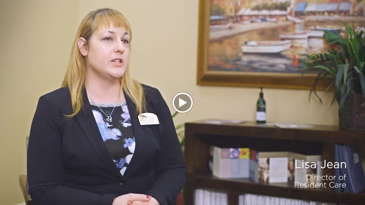 A woman with blonde hair wearing a black blazer and a floral top is seated indoors, speaking. Behind her is a wooden shelf with brochures and a bottle, a potted plant, and a framed painting of boats on water.