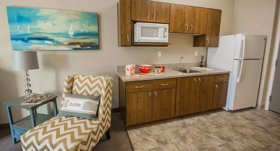 A small kitchenette area with wooden cabinets, a microwave, a sink, and a white refrigerator. To the left, there is a chevron-patterned armchair with a pillow that says 'home' and a small side table with a lamp and eyeglasses. A painting of sailboats on the water hangs on the wall above the chair.