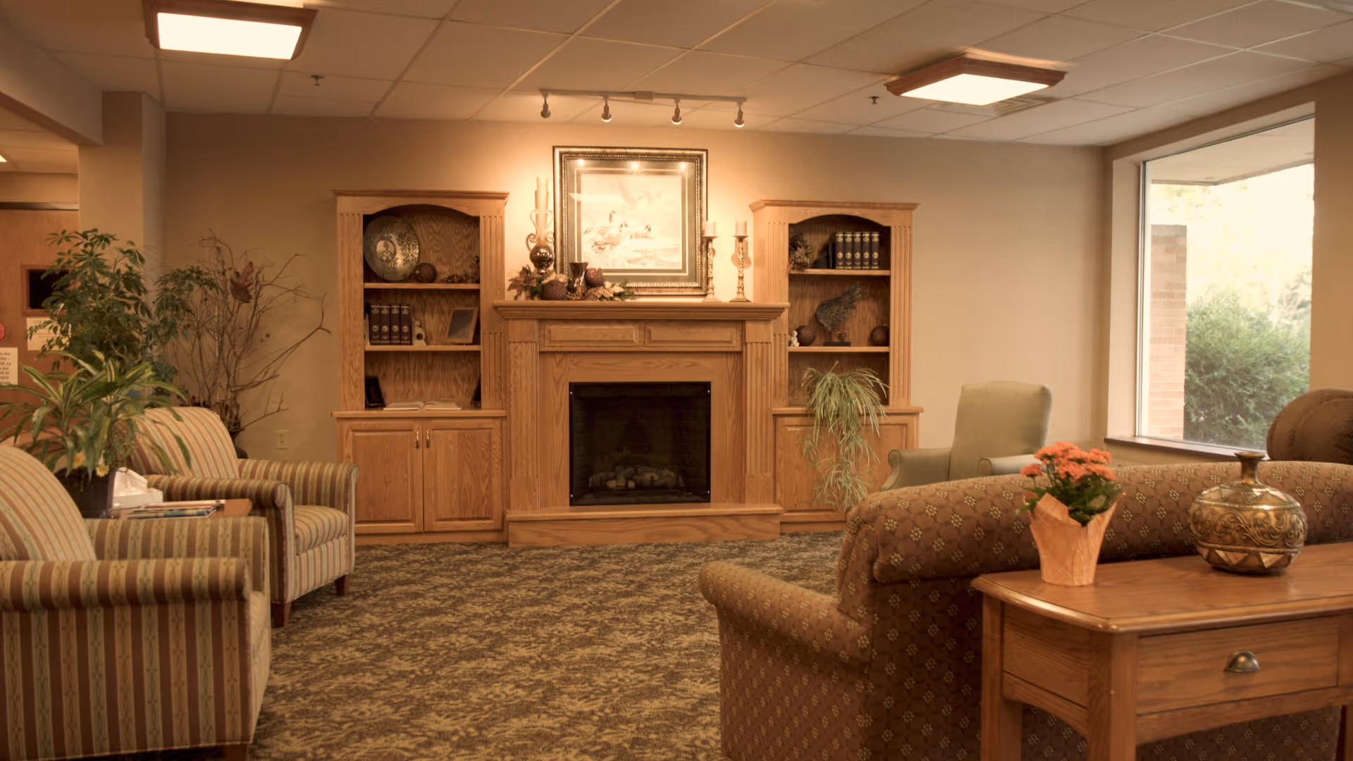 A cozy living room area with a central wooden fireplace flanked by two wooden bookshelves. The room features patterned carpet, several upholstered chairs and sofas, a wooden side table with a potted plant and decorative vase, and a large window letting in natural light.