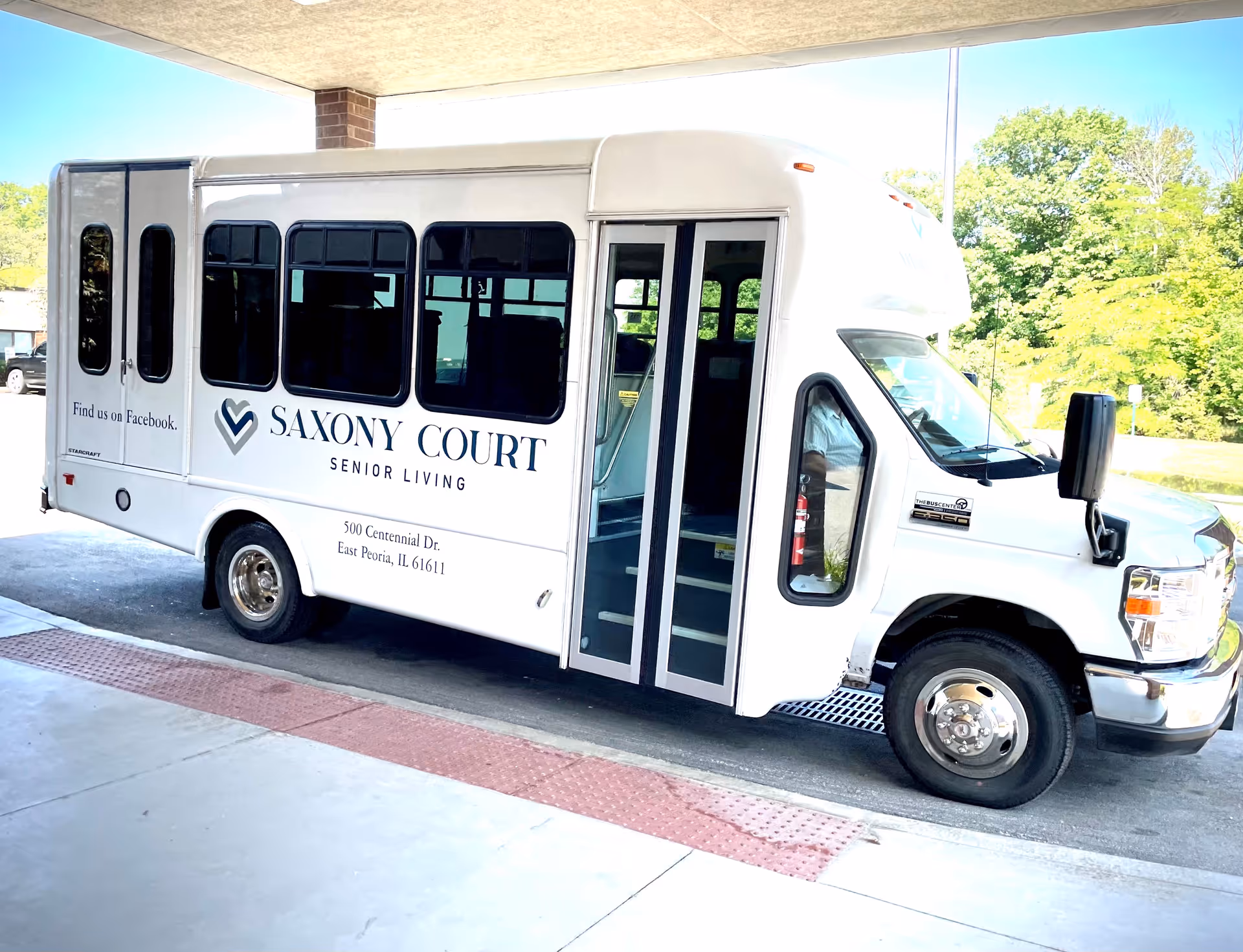 White Saxony Court Senior Living shuttle bus parked under a covered entrance with the facility name and address on its side.