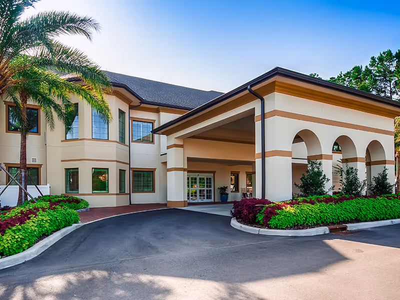 Entrance of a two-story senior living building with a covered porte-cochère, arched columns, palm trees and landscaped flower beds.