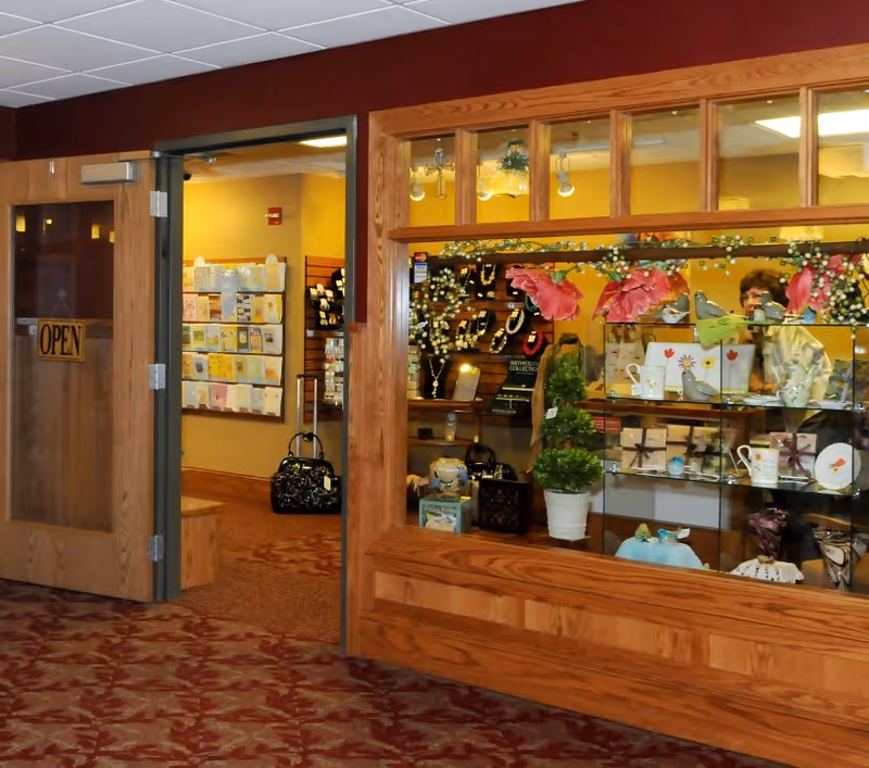Interior view of a gift shop area within a senior living facility. The shop has wooden framed glass display cases filled with various items such as jewelry, mugs, and decorative pieces. To the left, there is an open wooden door with an 'OPEN' sign hanging on it, leading to another room with greeting cards displayed on the wall. The floor is carpeted with a red and brown patterned design.