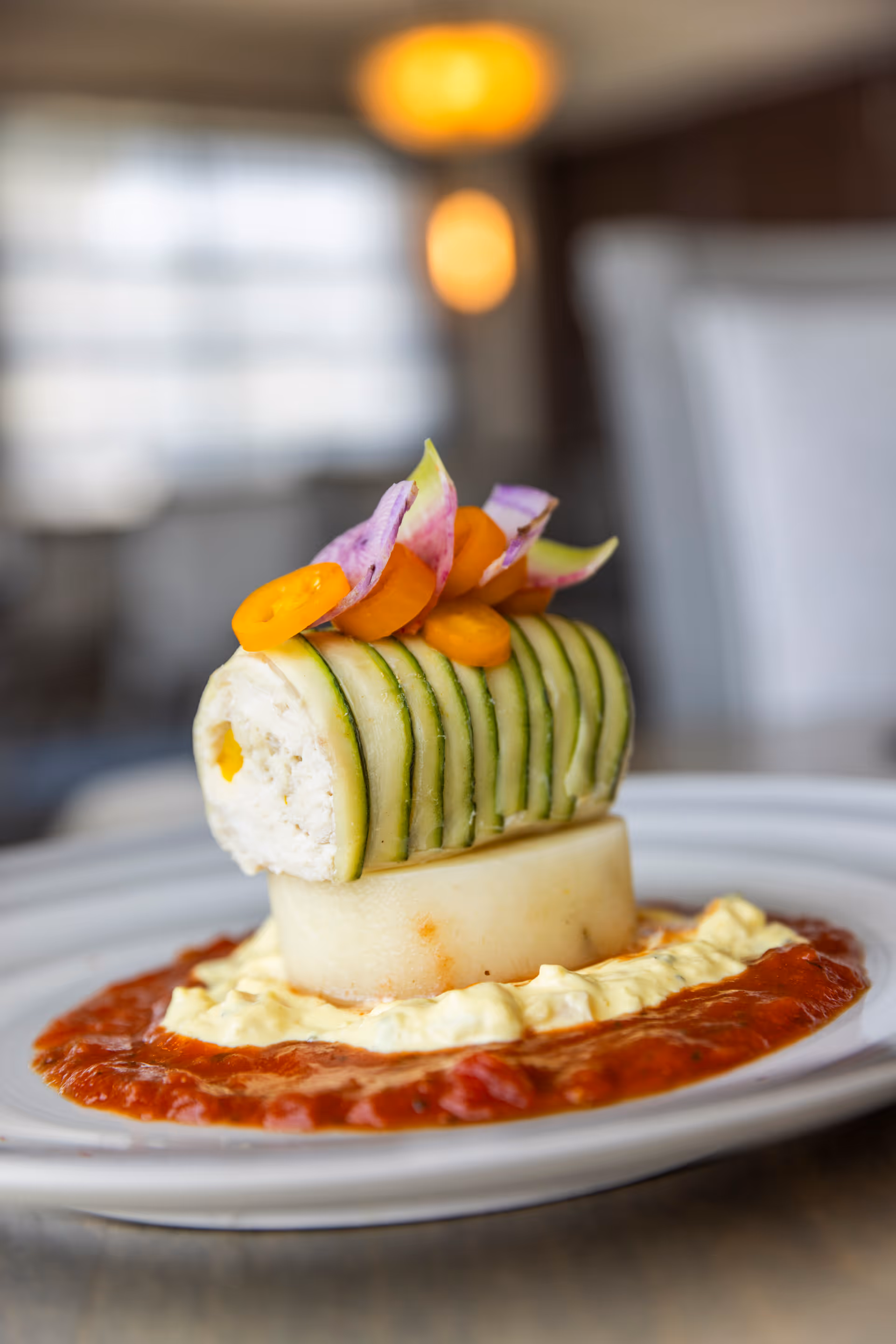 A gourmet plated dish featuring a rolled cucumber slice filled with a creamy mixture, garnished with thin carrot slices and purple flower petals, served on a white plate with a base of red sauce and a creamy yellow sauce.