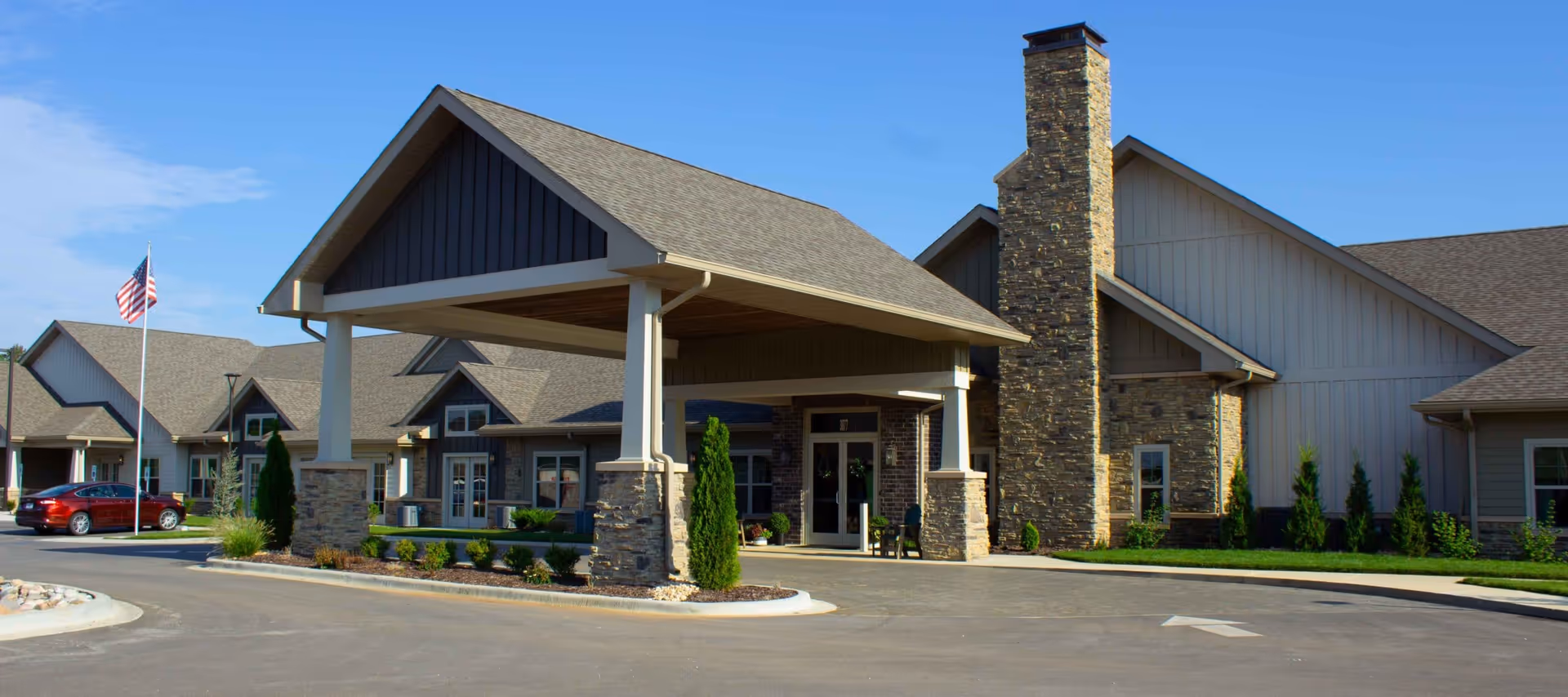 Front exterior of a single-story senior living facility with a covered porte-cochere, stone chimney, and driveway.