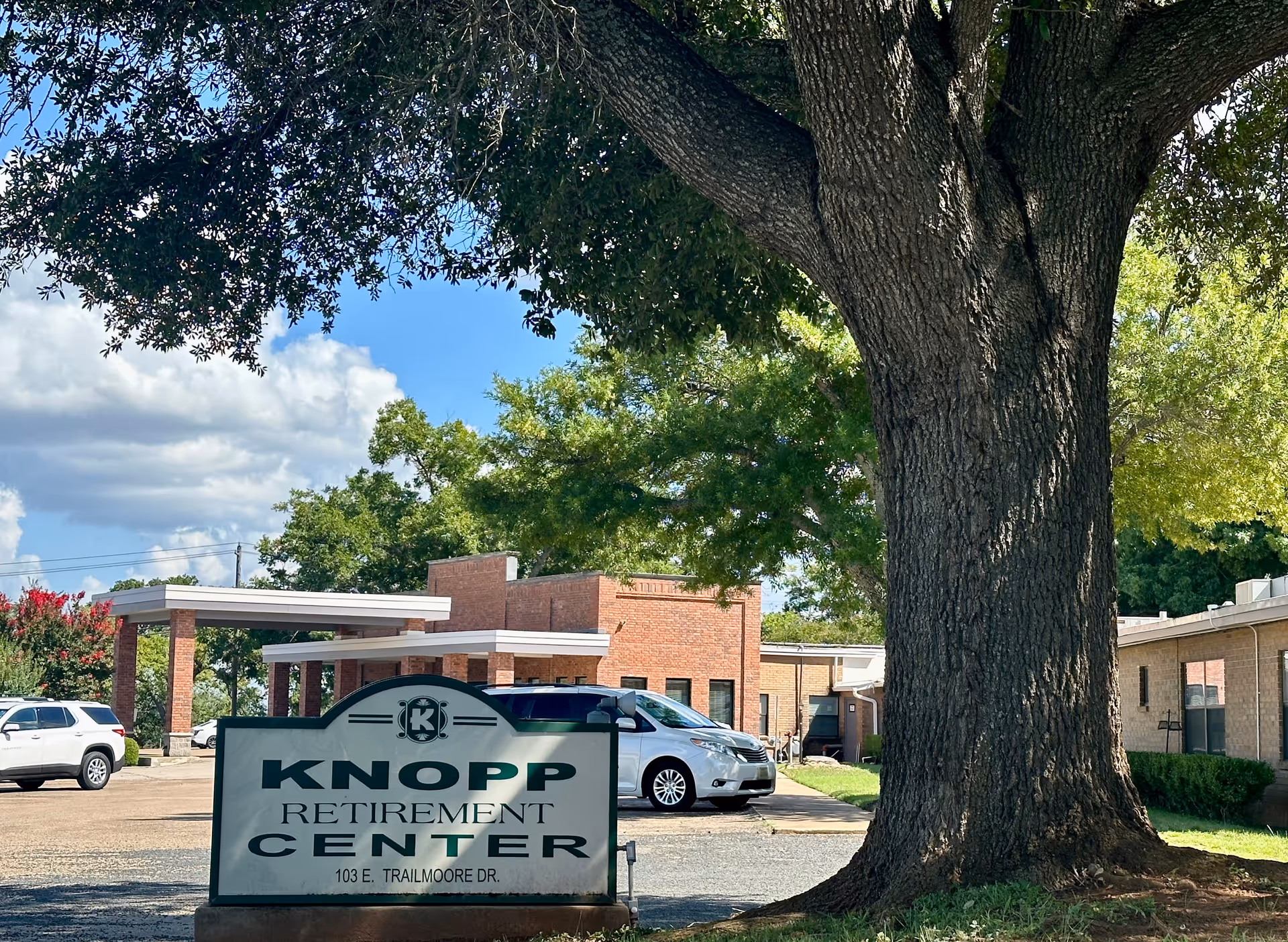 Exterior view of Knopp Retirement Center showing a large tree in the foreground, a sign with the facility's name and address, several parked cars, and a brick building under a partly cloudy blue sky.