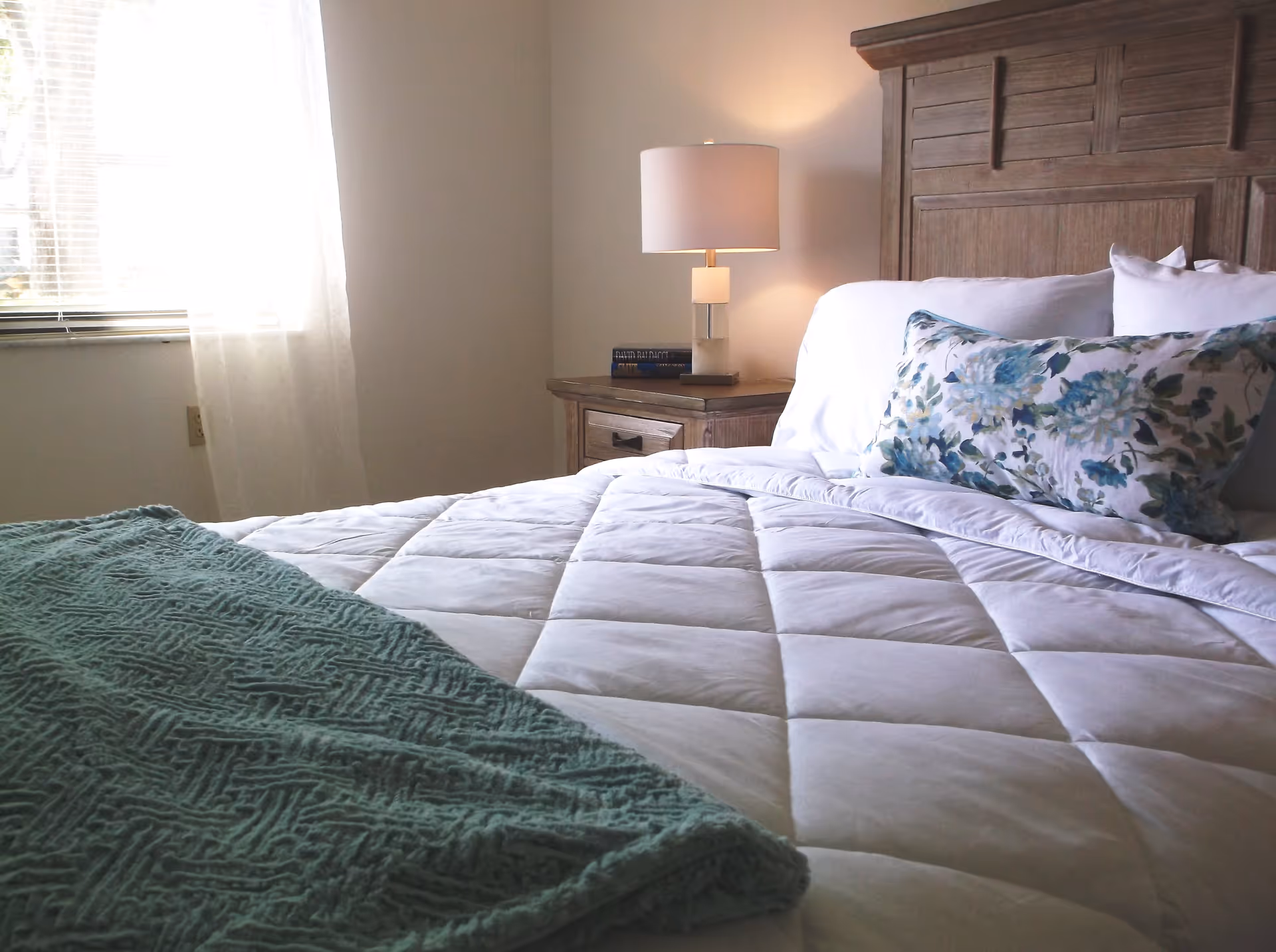 Sunlit bedroom with a made bed, floral pillow, wooden headboard and a bedside table with a lamp.