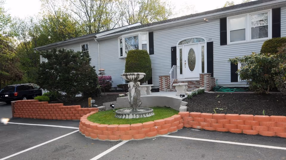 Front exterior of a single-story residential building with a decorative fountain, brick flowerbed borders, and parking spaces in front.